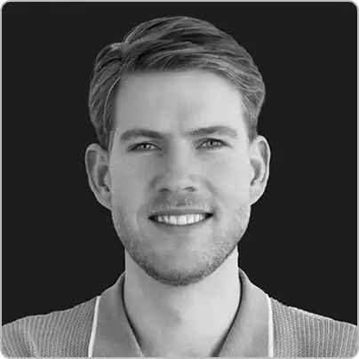 About Us: Portrait of a young man with short hair and light stubble, smiling, wearing a collared shirt, and facing forward against a plain dark background.