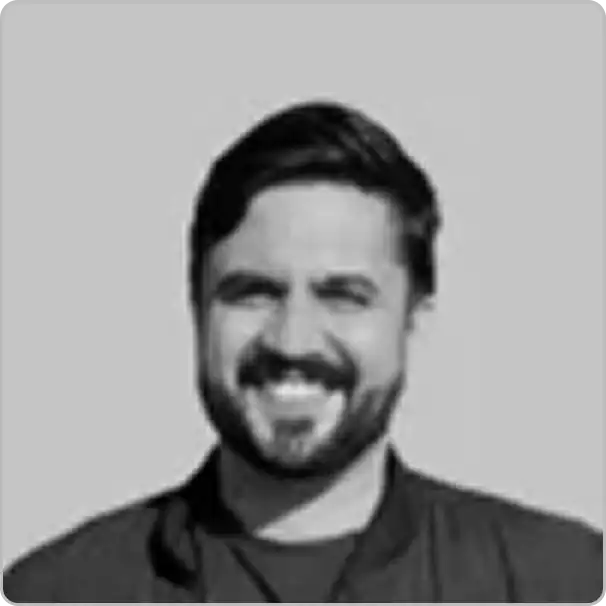 A man with dark hair and a beard, smiling, is shown from the shoulders up in a black and white photo with a plain background—an inviting portrait perfect for an About Us page.