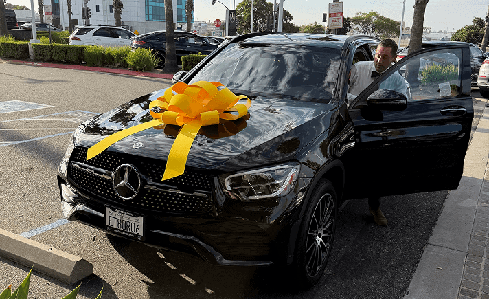 A man stands by an open door of a black Mercedes-Benz SUV with a large yellow bow on the hood in a parking lot.