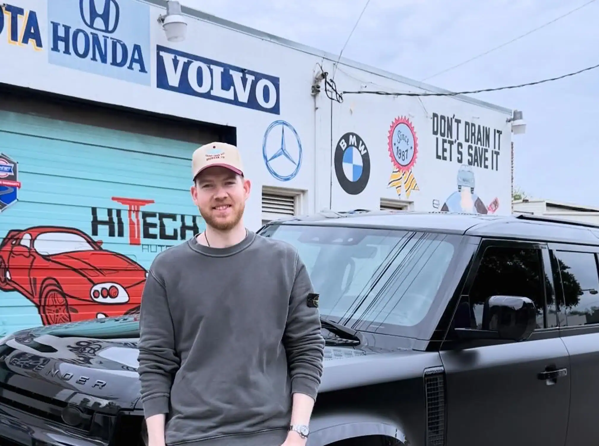 A man in a gray sweatshirt and tan cap stands in front of a black SUV, with an auto shop featuring car brand logos and murals in the background—capturing the spirit of our About Us story.