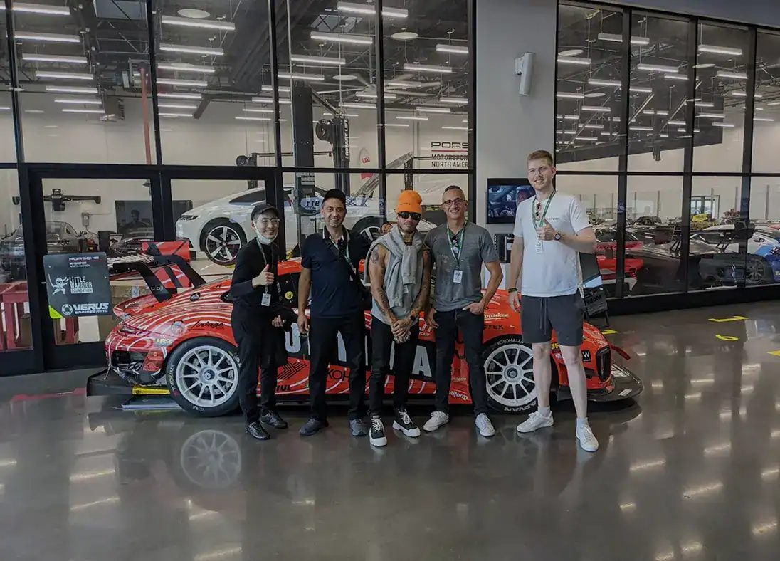 Five people stand in front of a red race car inside a showroom with cars visible behind glass walls, capturing the spirit of our About Us story.