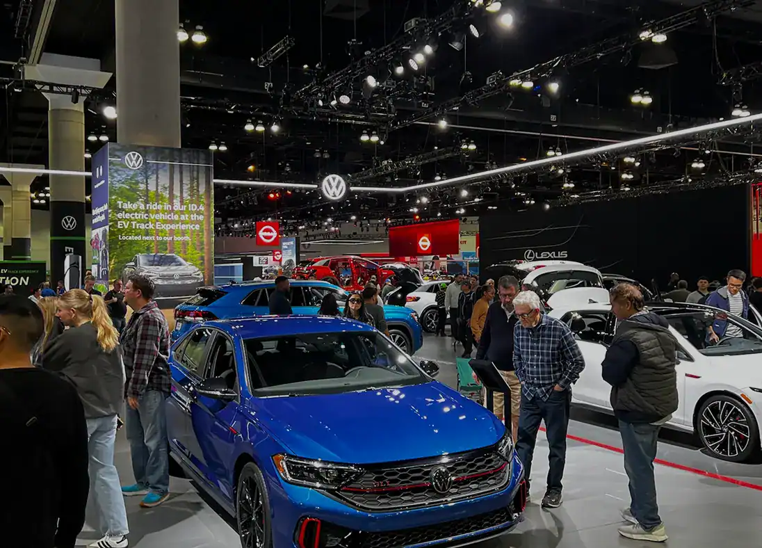 People view Volkswagen cars at an indoor auto show with various vehicles on display and branded signs in the background, offering a unique glimpse into what makes Volkswagen special—much like learning more in the About Us section.