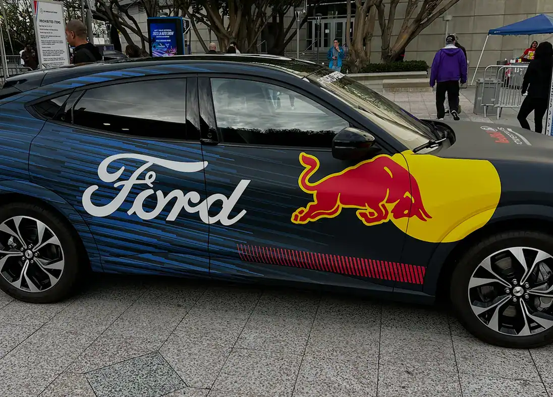 A car with Ford and Red Bull logos is parked on a paved area outside a building, with people walking in the background, offering a dynamic glimpse into our brand’s presence—learn more on our About Us page.