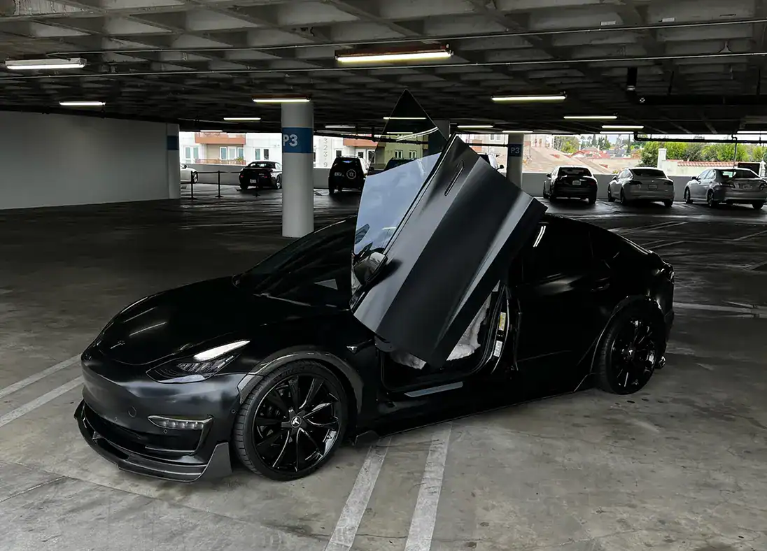 A black Tesla Model 3 with vertical doors open is parked in a multi-level parking garage, perfectly capturing the innovative spirit found in our About Us story.