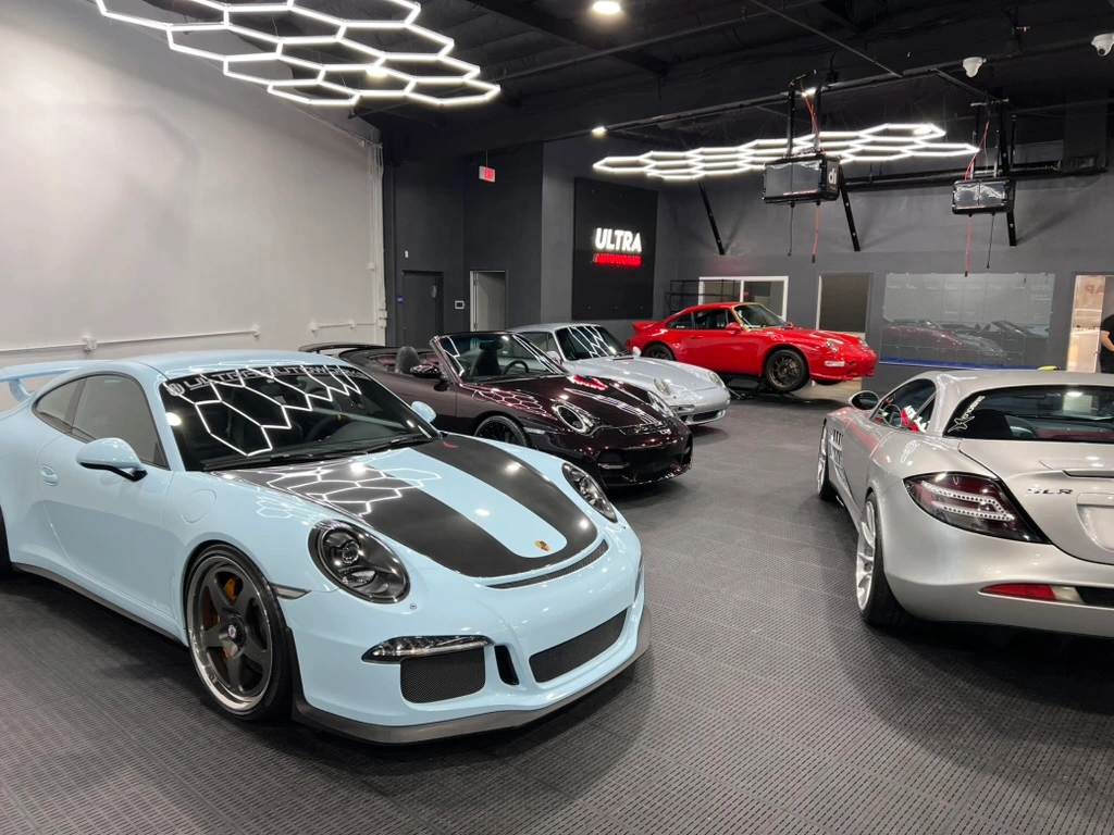 Several high-end sports cars, including Porsches and a Mercedes, are parked inside a modern, well-lit showroom with black walls and geometric ceiling lights.