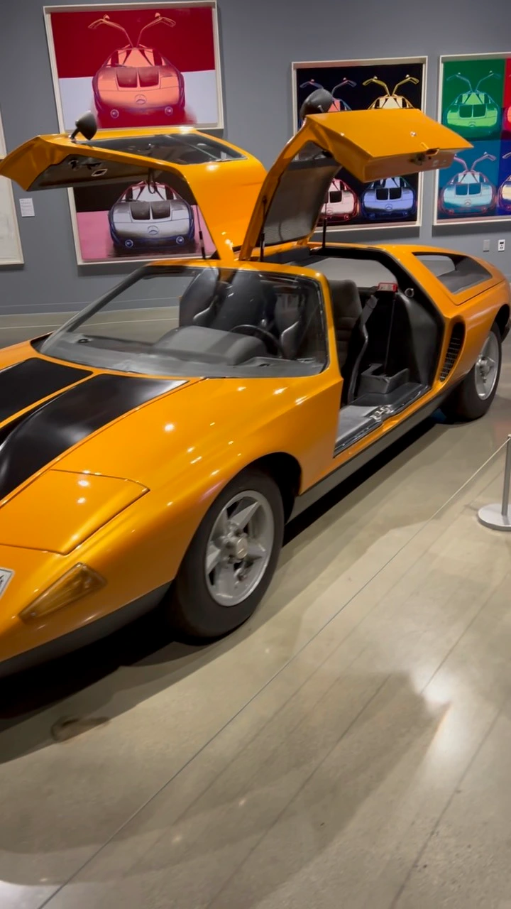 Yellow sports car with gullwing doors open is displayed indoors next to colorful car art on the wall.