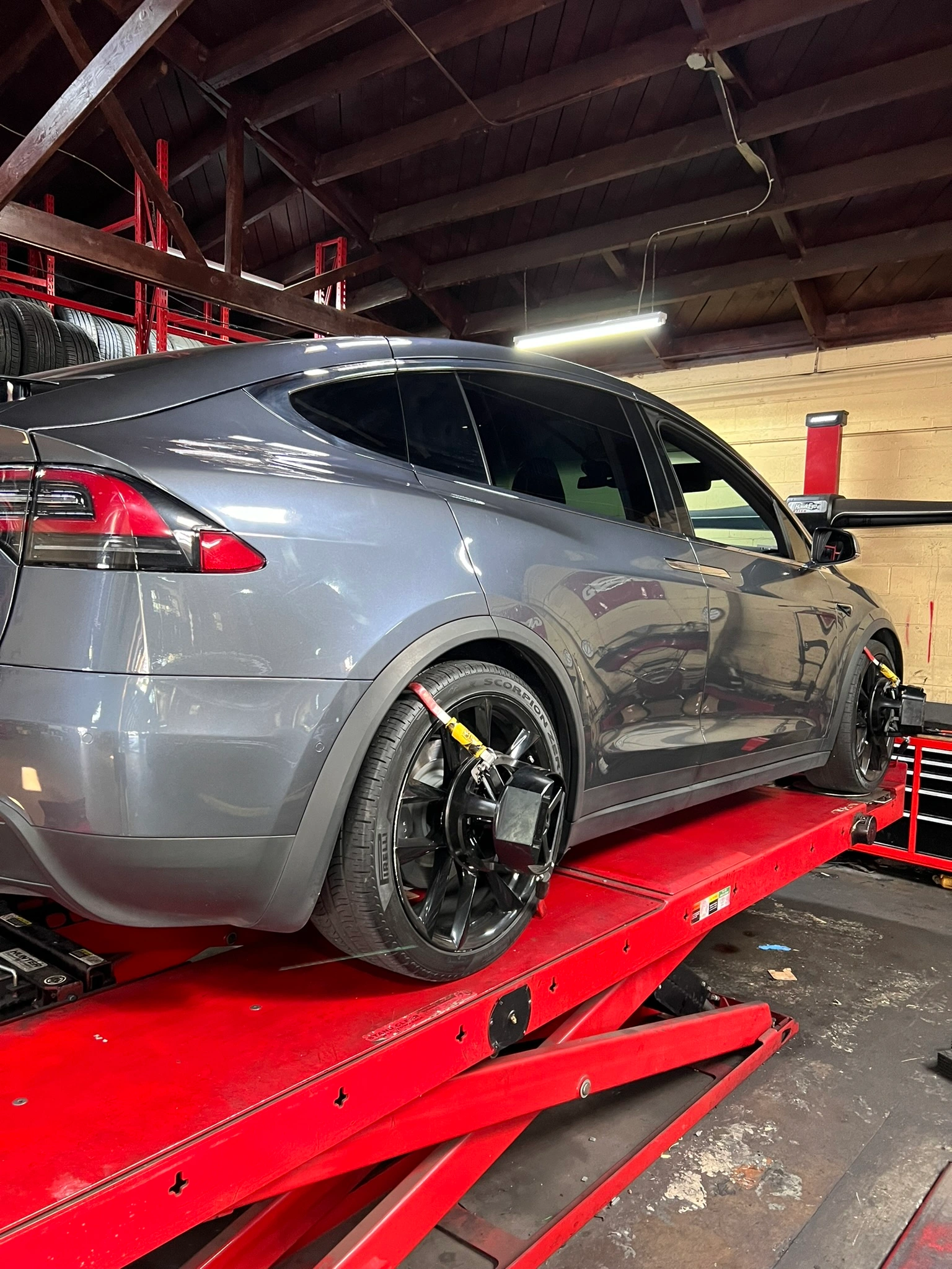 A gray Tesla SUV on a red hydraulic lift undergoing a wheel alignment in an auto repair shop with various tools visible.